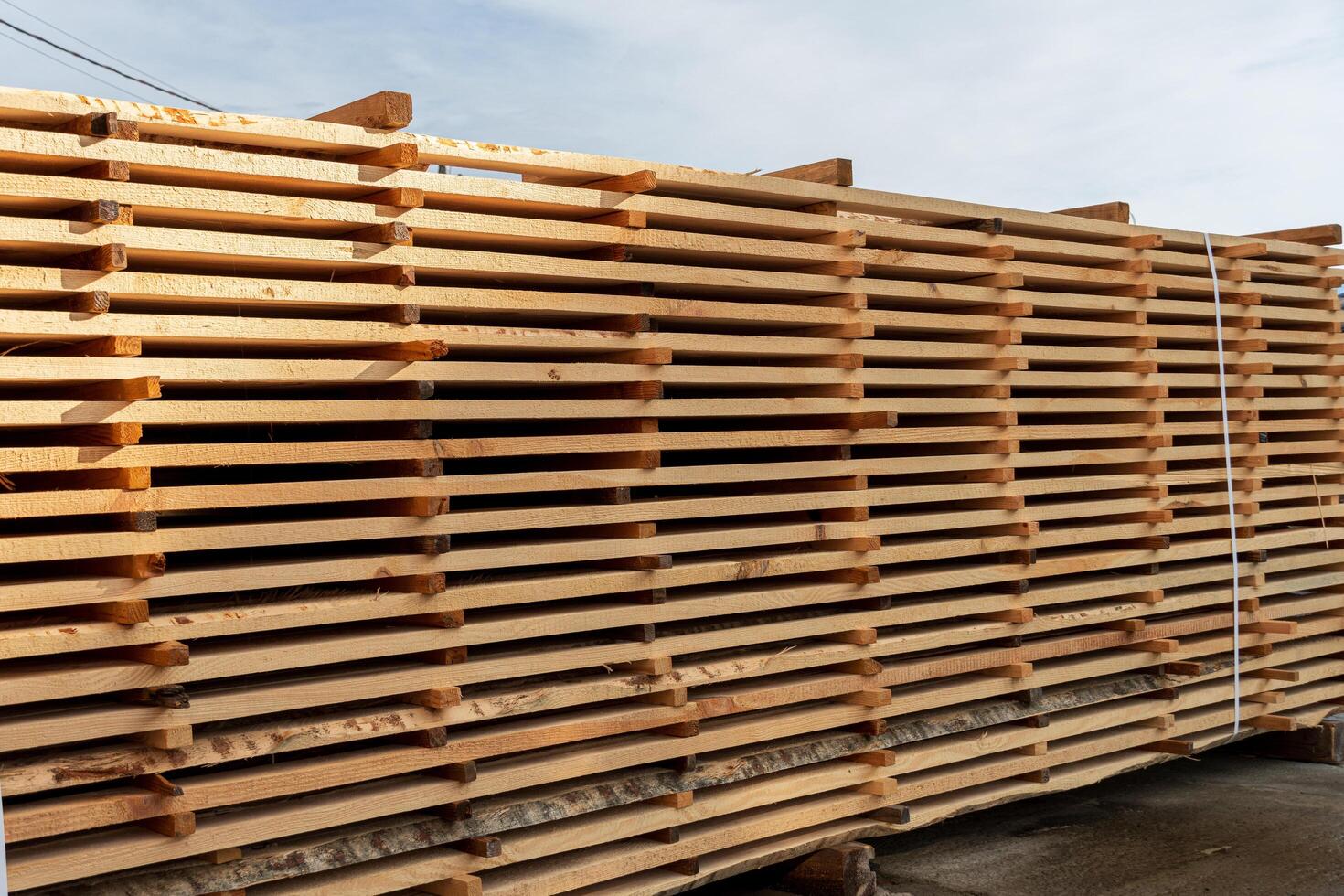 Stacks of freshly cut wooden planks are neatly arranged at a lumber yard, illuminated by afternoon sunlight against a clear sky photo