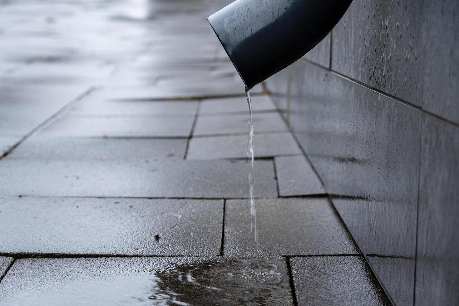 Water drips steadily from a drainpipe onto a smooth, wet pavement after recent rain, creating puddles on the ground photo