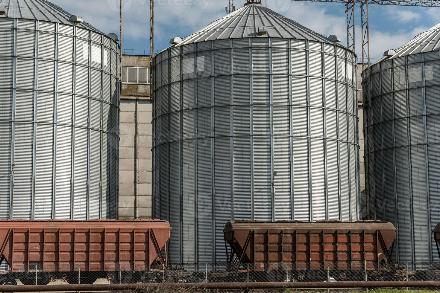 Grain is being stored in large silos at an agricultural site, with train cars ready for transportation in the background photo