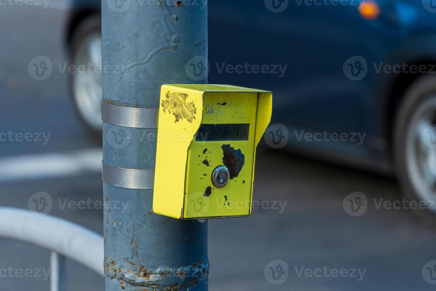 A weathered pole supports a bright yellow traffic signal button, with a car driving past on a bustling urban street photo