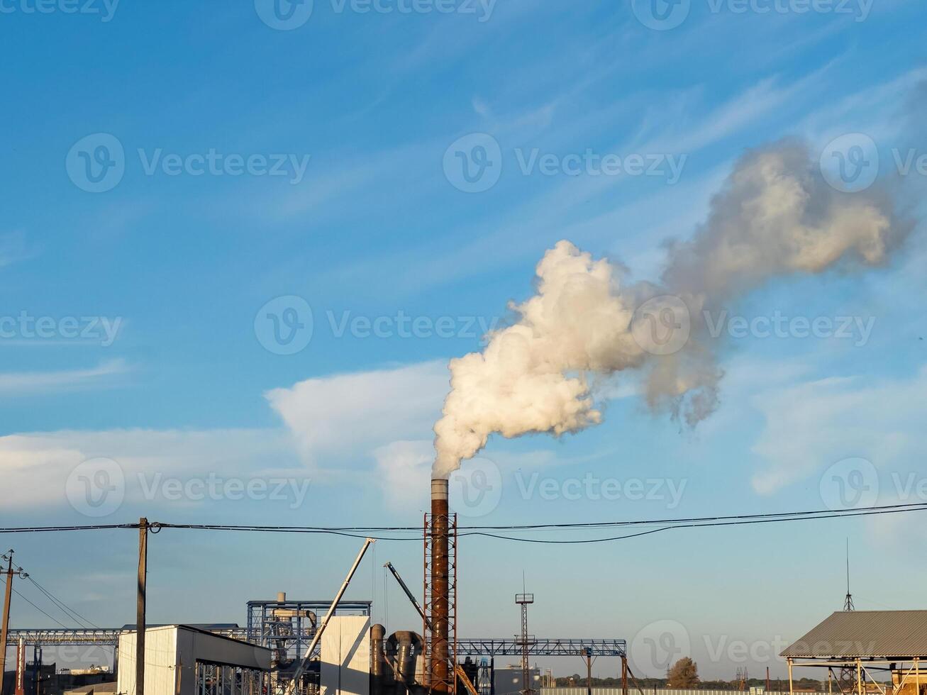 A large factory releases white smoke from its chimney against a backdrop of a clear blue sky during late afternoon hours photo