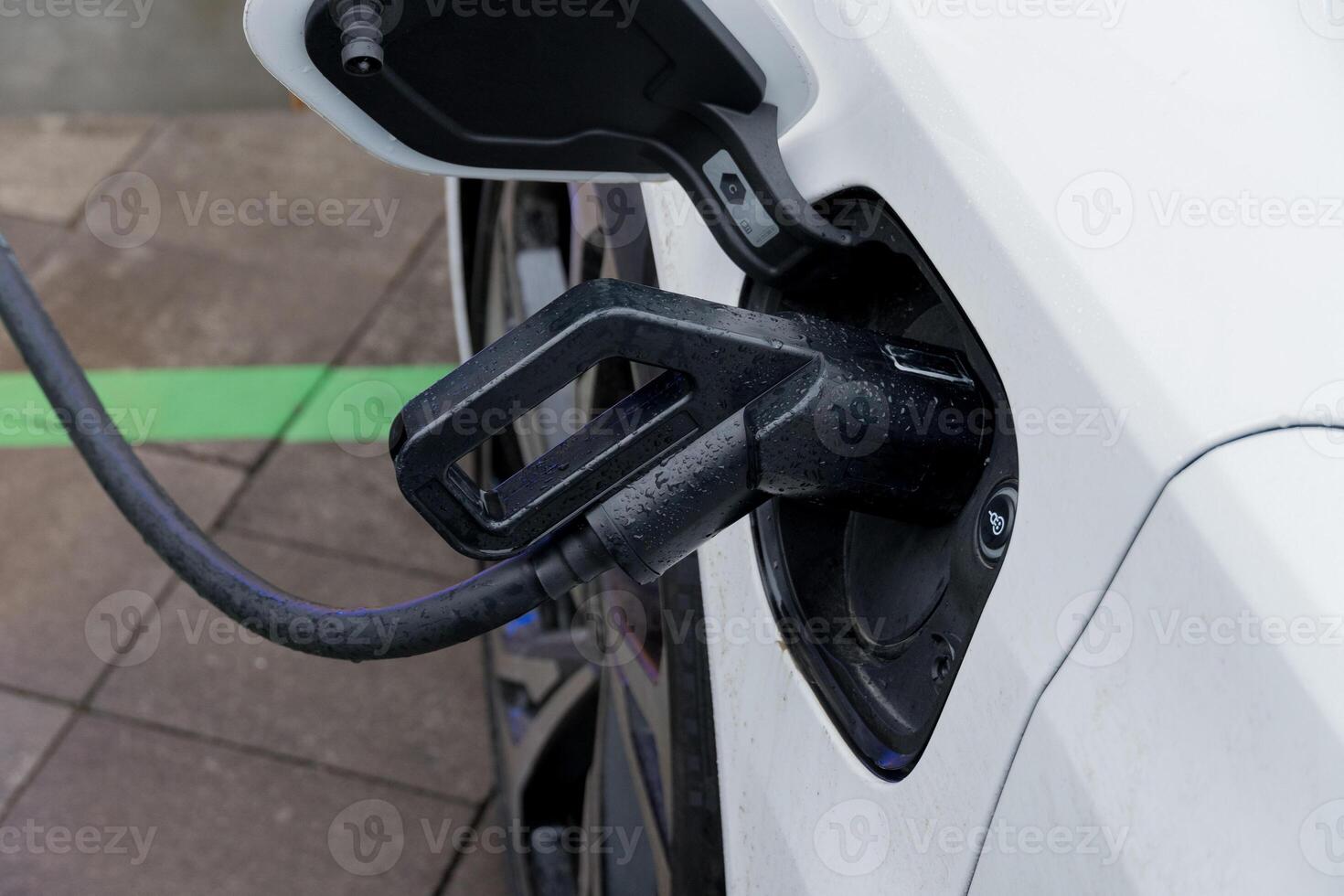 A white electric vehicle is connected to a charging station, with the charging cable securely plugged in, capturing the urban atmosphere photo