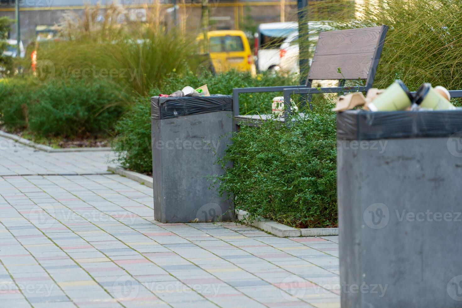 Littered waste bins sit by a park bench, surrounded by greenery and paved stones in a busy urban environment photo
