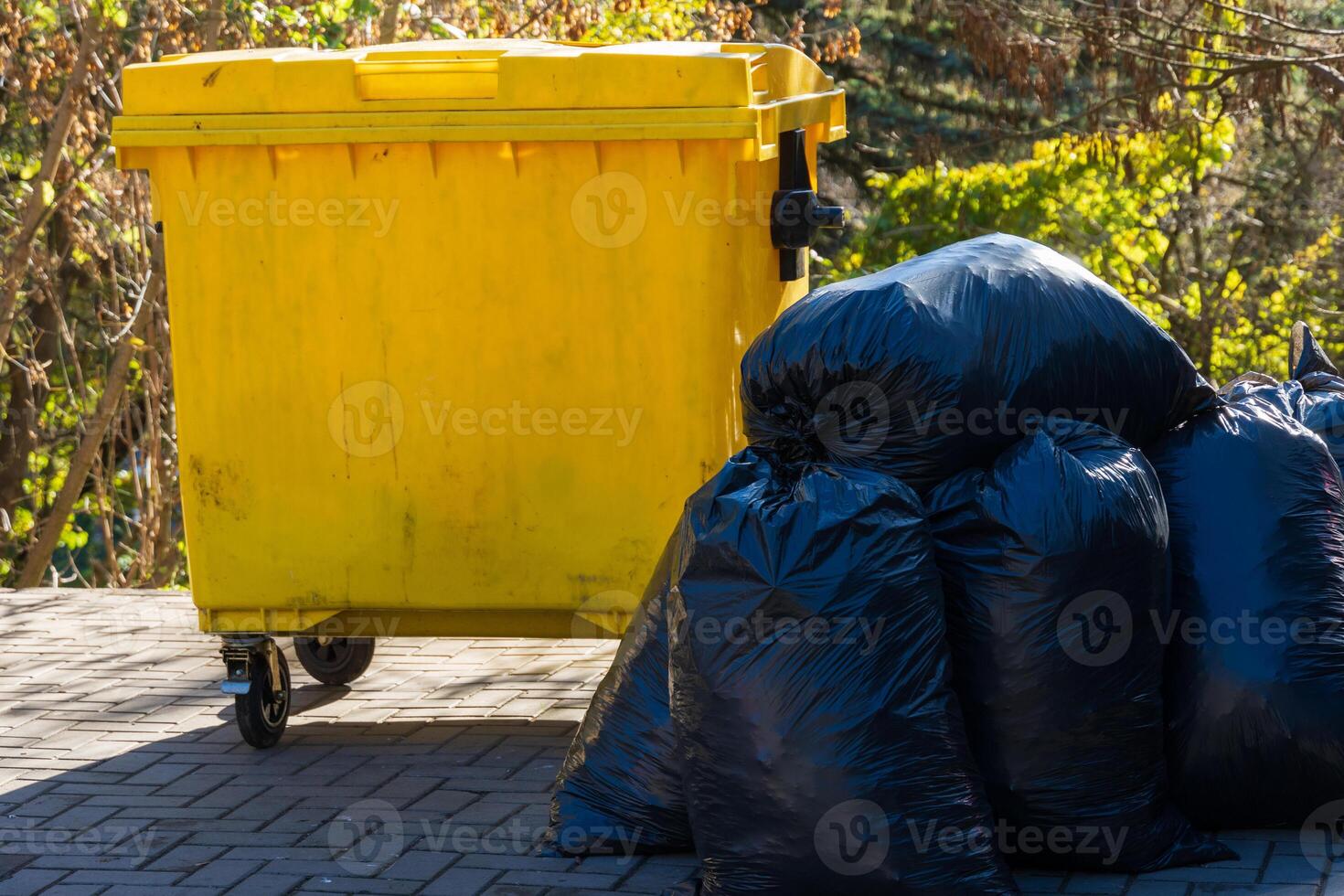 A yellow dumpster is positioned next to several black garbage bags in a tidy outdoor environment during daylight hours photo