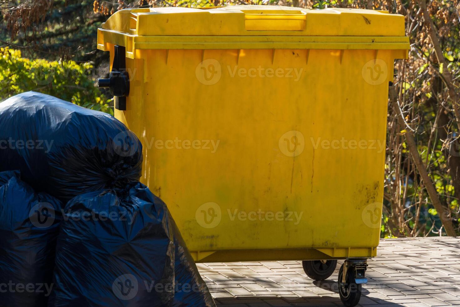 A bright yellow waste container is positioned next to several black garbage bags in a park setting during daylight hours photo