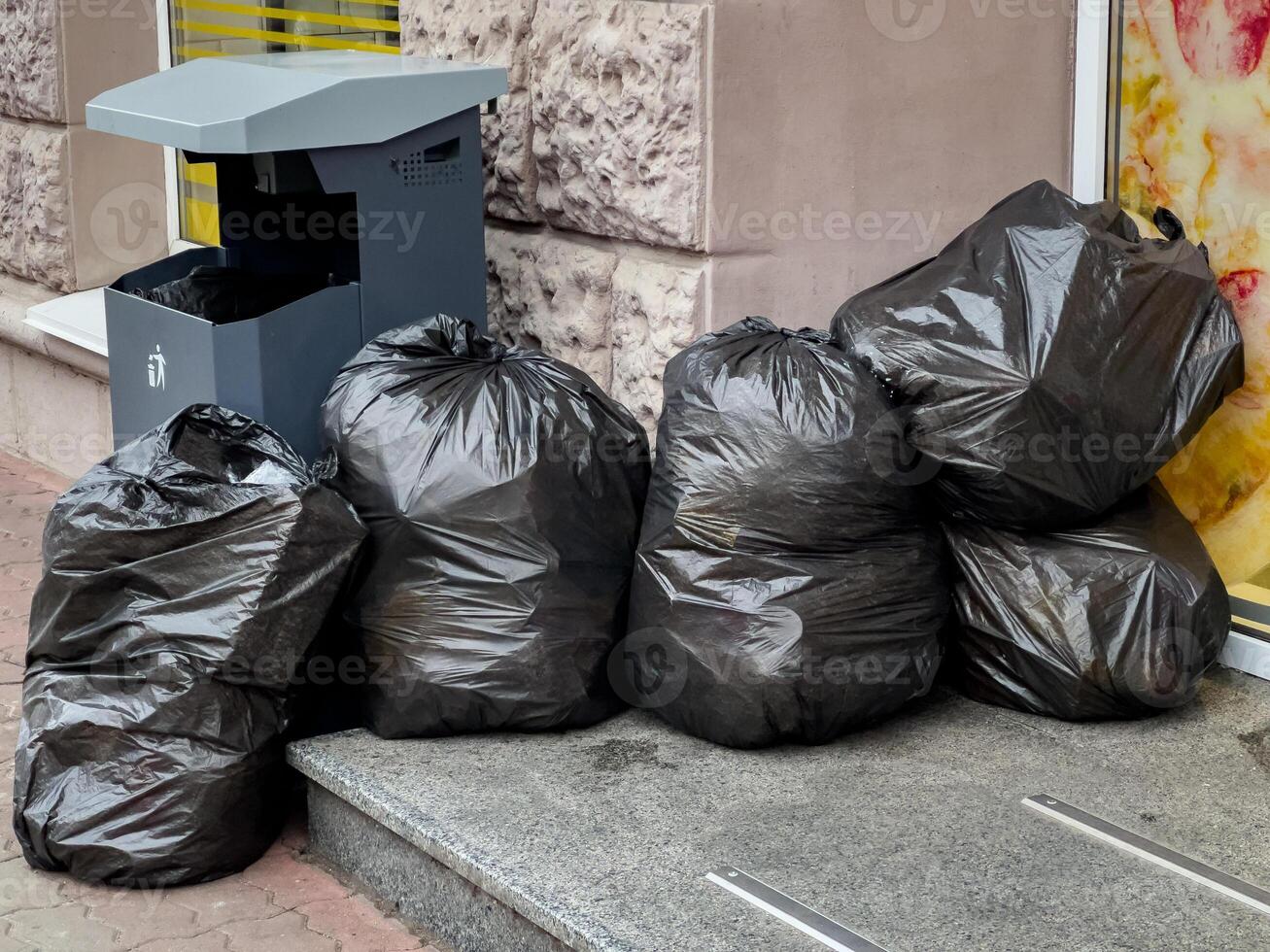 Multiple black trash bags are piled on the sidewalk next to a litter bin in a city setting photo