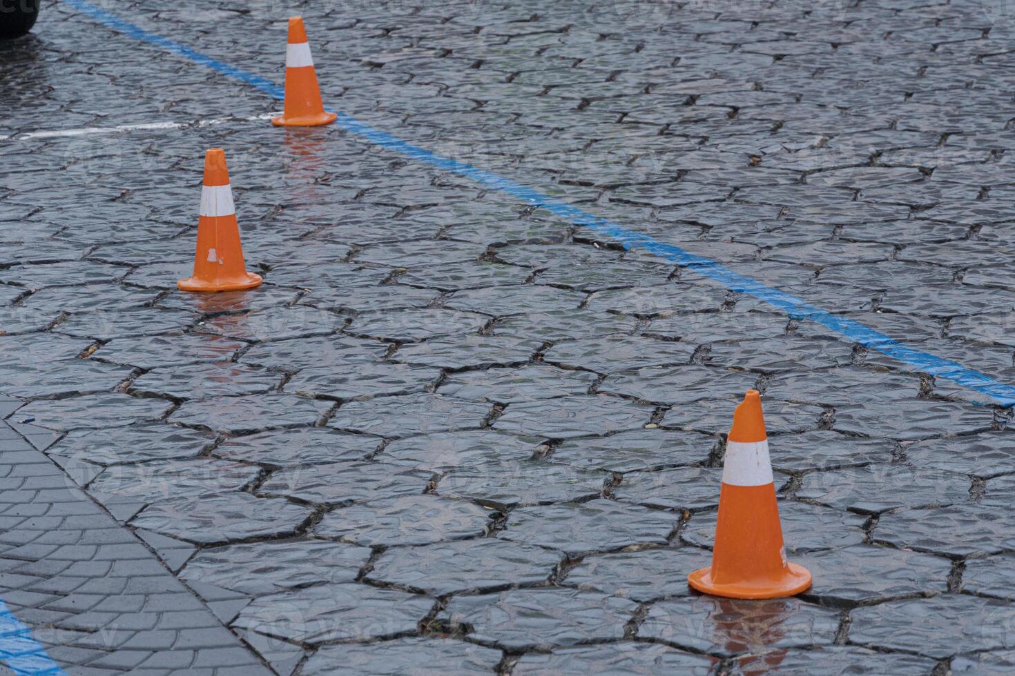 Orange traffic cones sit on a wet cobblestone surface, marking no parking zones and creating safety measures in rainy conditions photo