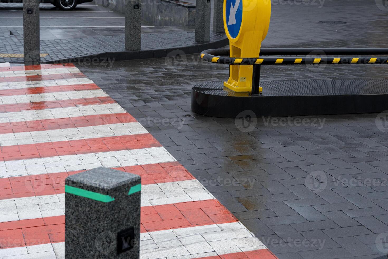 A crosswalk featuring red and white stripes leads to a yellow warning post in a modern urban area on a rainy day photo