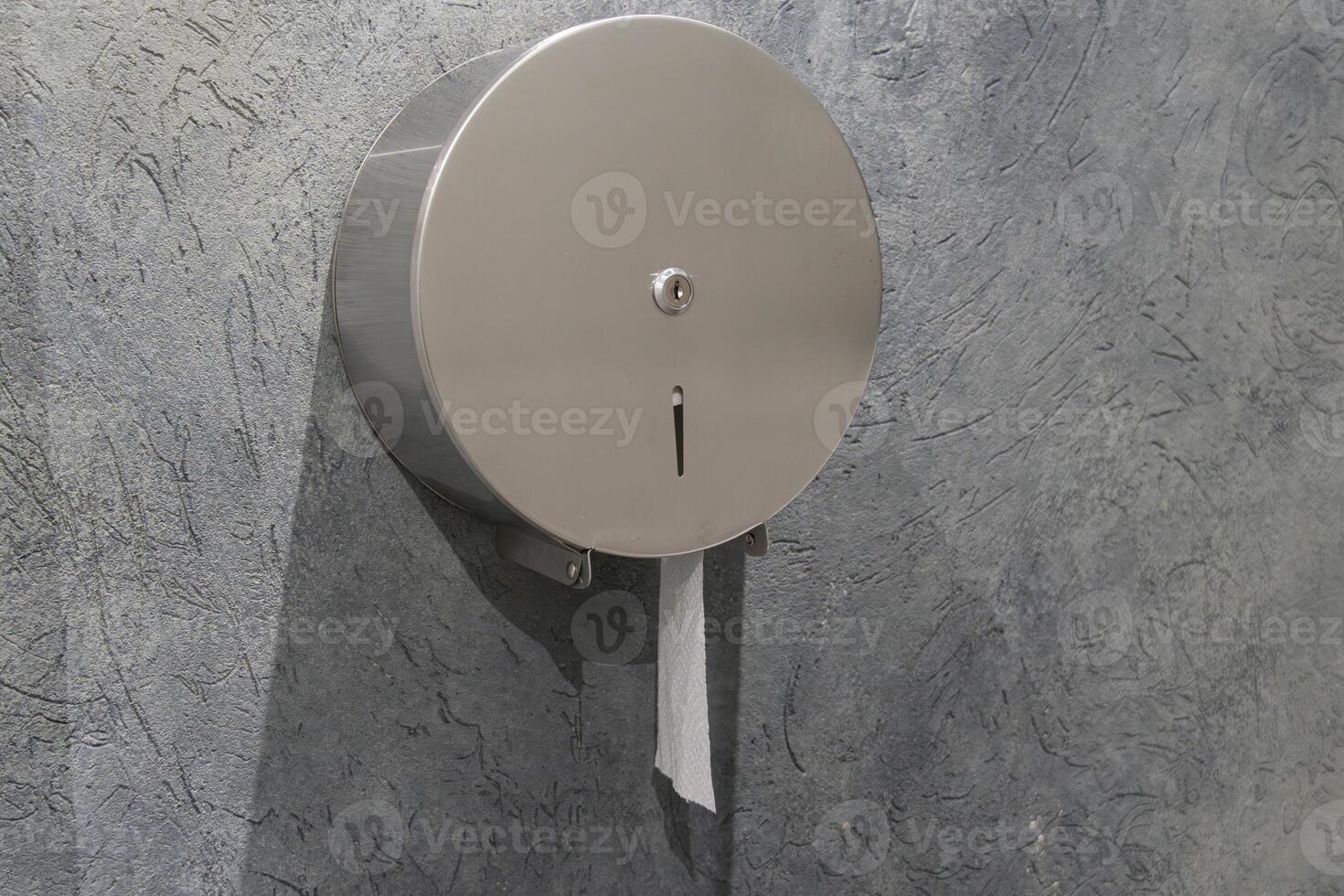 A stainless steel tissue dispenser is securely mounted to a textured gray wall in a public restroom, providing easy access for users photo