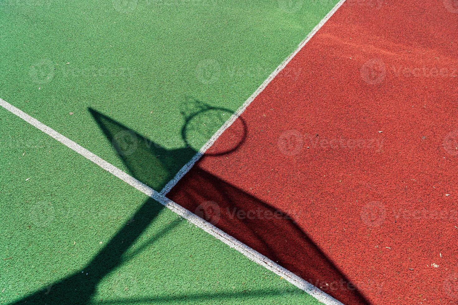 A colorful basketball court in bright sunlight shows a hoop's shadow stretching across red and green surfaces, inviting joyful games photo