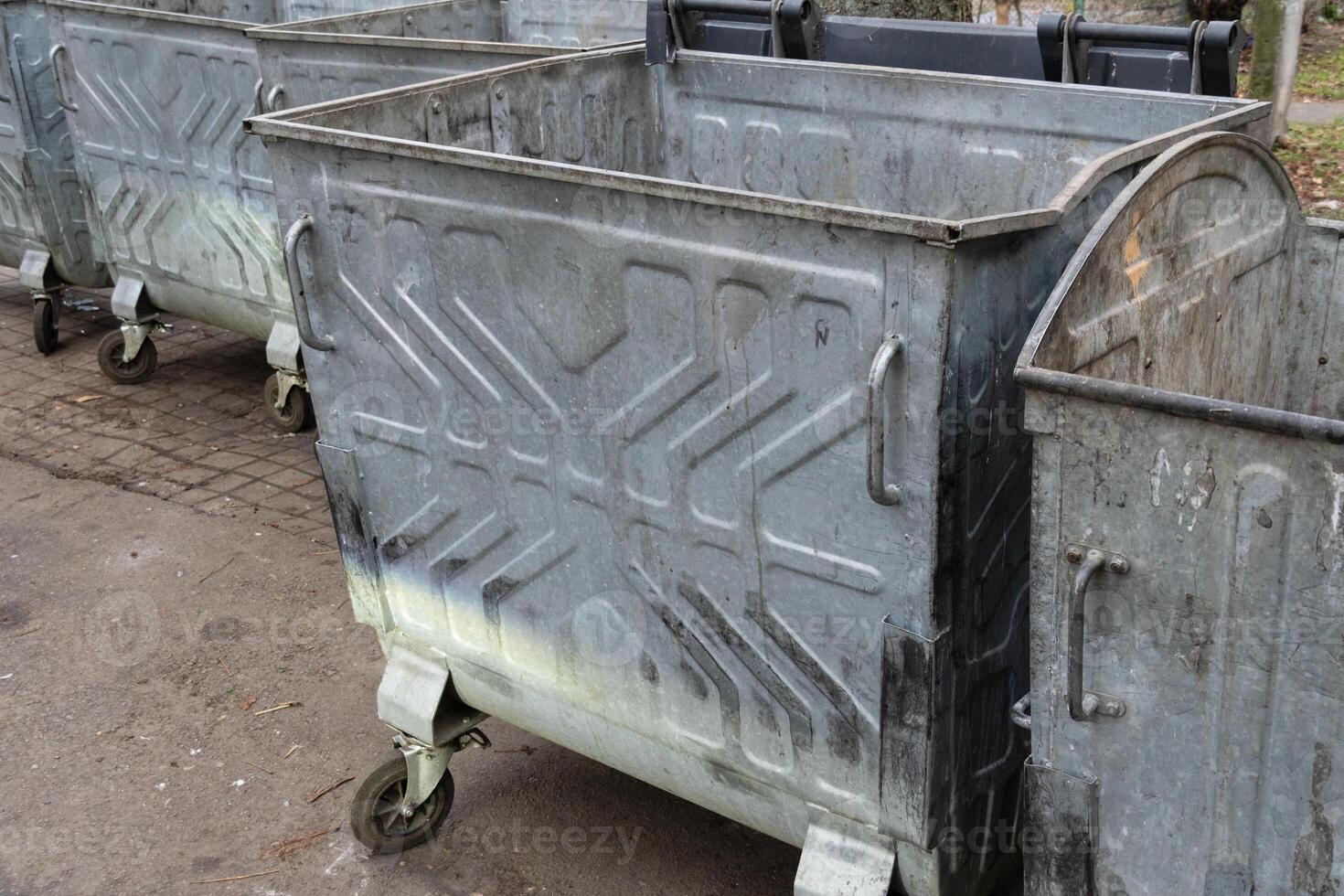 Several metal dumpsters are positioned in a city area, showing signs of wear and tear, with a clear sky overhead photo