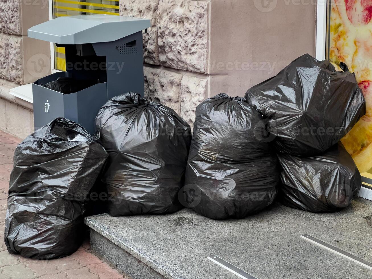 Black trash bags rest against a wall next to a recycling bin, emphasizing the need for proper waste disposal in city spaces photo