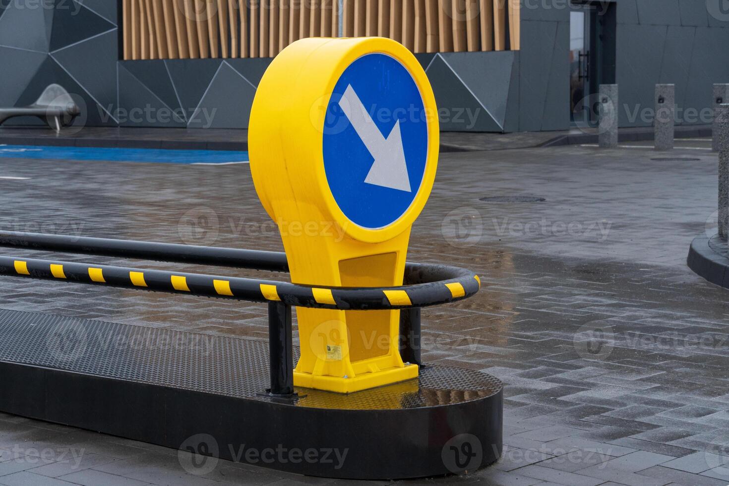 In an urban plaza, a vibrant yellow sign directs foot traffic with its clear arrow design amidst modern structures and wet pavement photo