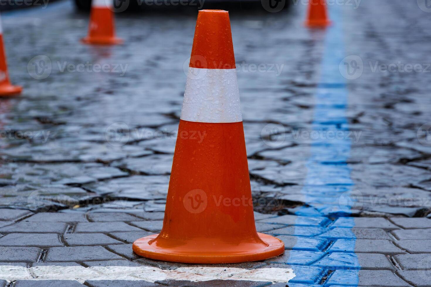 Rain-soaked cobblestone street features bright orange traffic cones marking a construction zone for pedestrian safety in an urban area photo