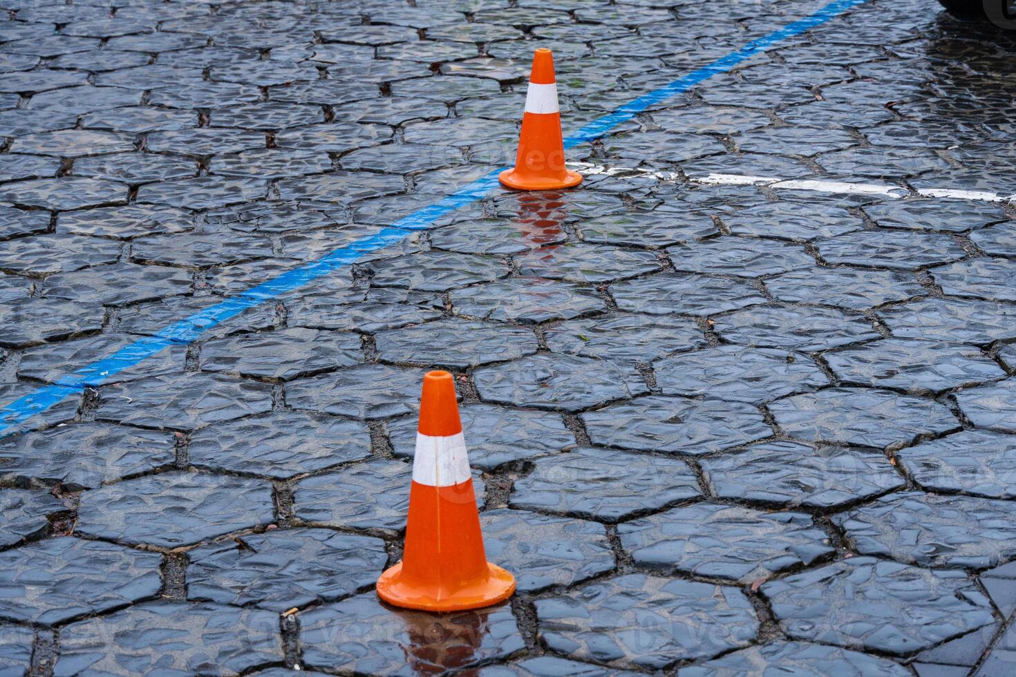 Traffic cones are positioned on a wet cobblestone street marking a construction zone in an urban area during rain photo