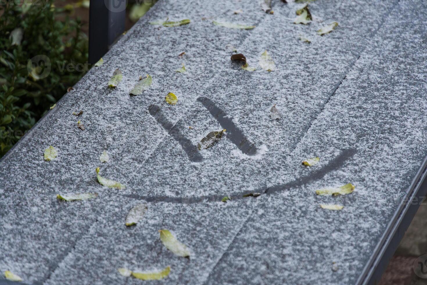 A bench in a winter landscape is blanketed with snow and frost, where a smiley face is playfully etched into the white layer photo