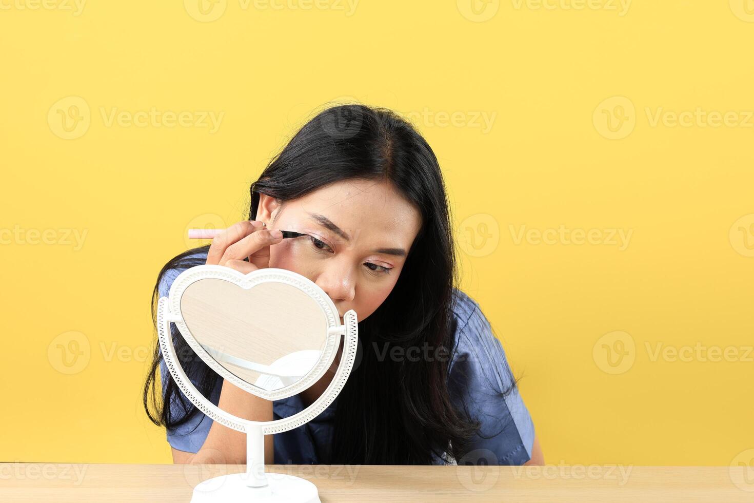 Young Asian Woman Applying Make-up to Her Face, using Black Eyeliner for Eyes and Looking at the Mini Standing Mirror photo