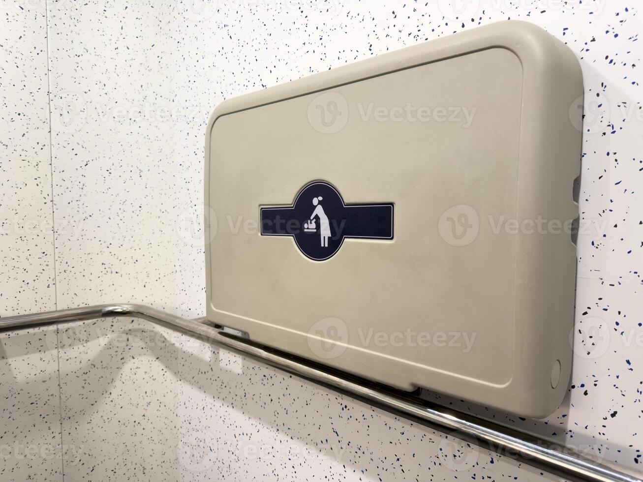 Wall mounted baby changing table in a public restroom with a stainless steel safety bar. Modern accessible facility for parents with infants, promoting convenience and inclusive design photo