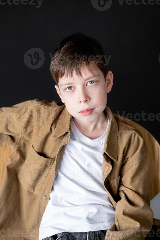 12 year old teenage boy with green eyes in brown shirt sits on chair against black wall and looks at camera with serious expression photo