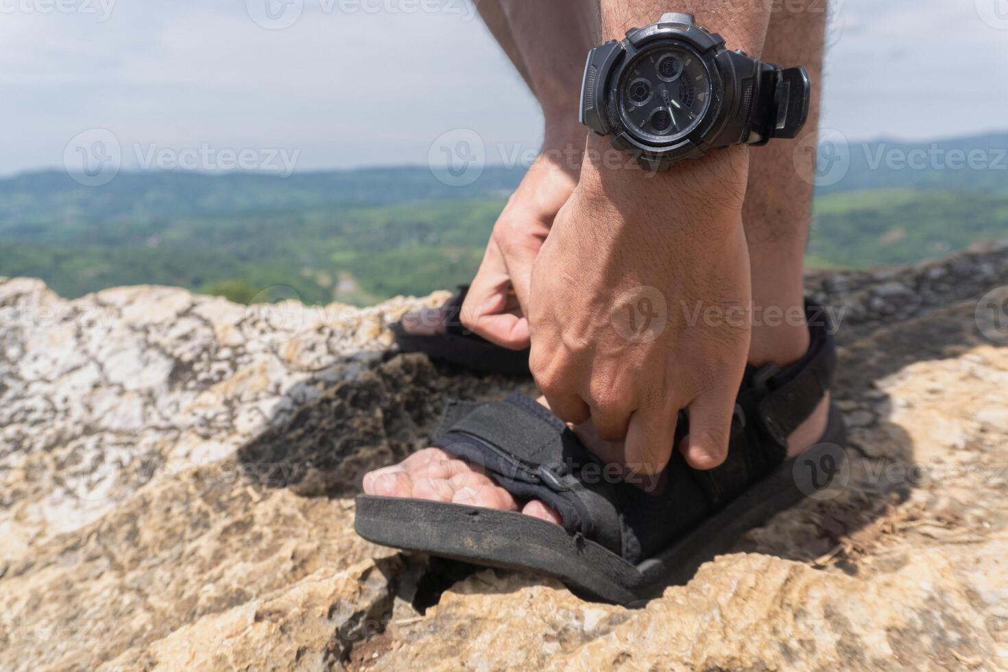 A person is adjusting their sandals outdoors on a rocky surface, with a watch visible on their wrist. photo