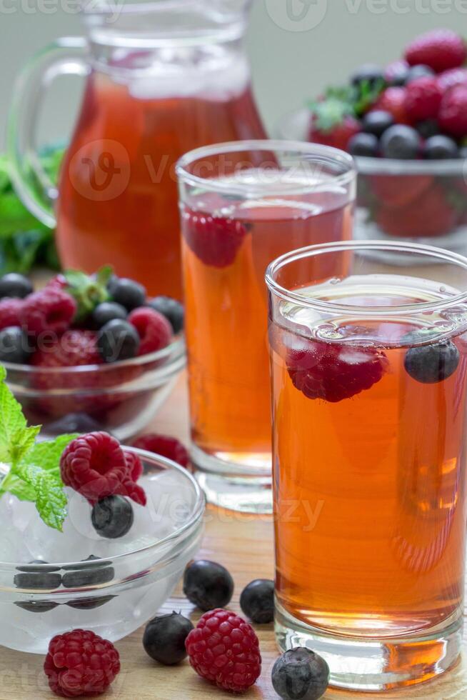 2 compotes of raspberries, strawberries, blueberries, near bowl of ice, 2 bowls of berries, mint leaves, carafe compote, straws on light background scattered berries. Berries compote. Vertical. Close. photo
