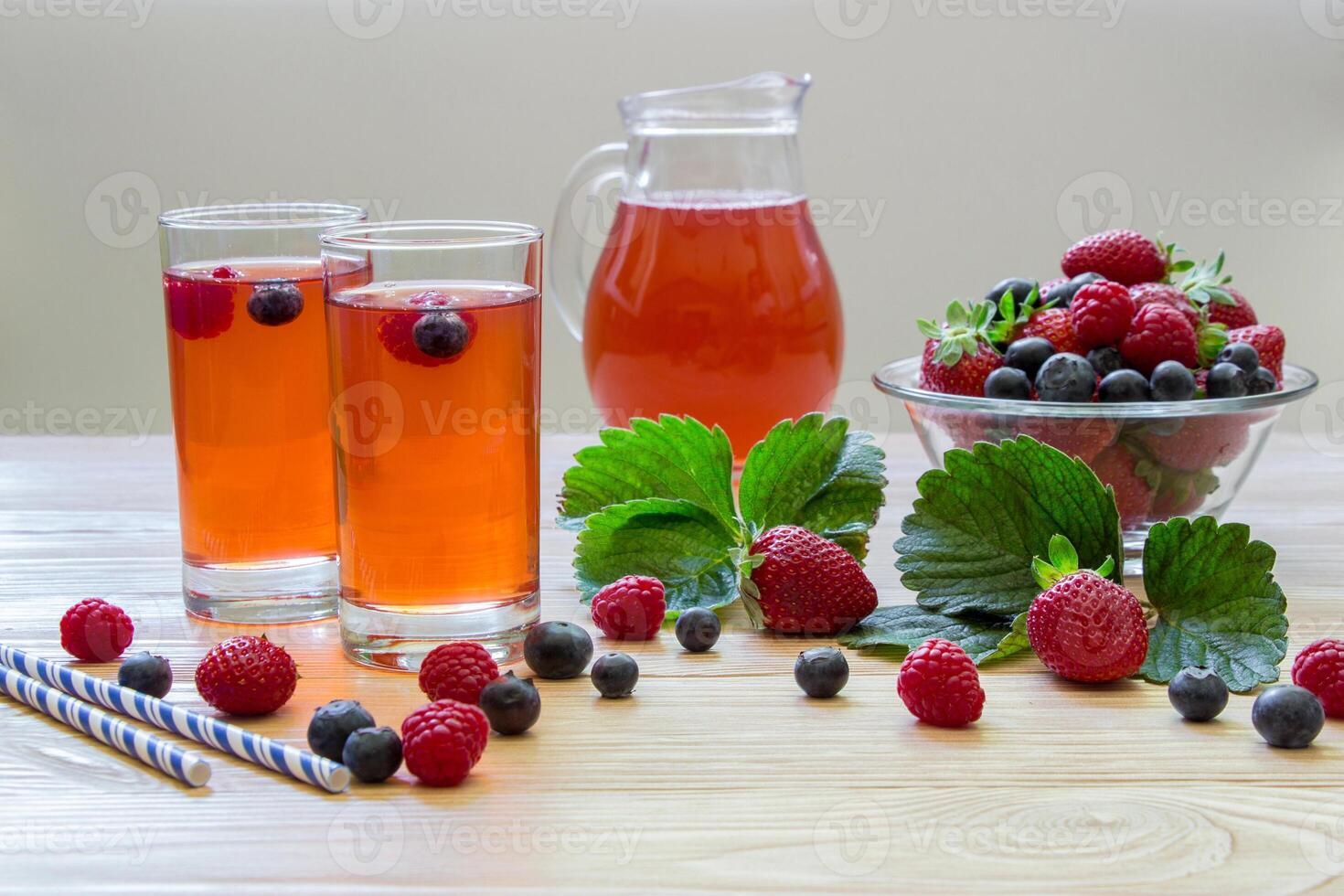 Left two glasses of berries compote, near bowl with berries, carafe compote on a light wooden background scattered berries and some empty space . Fresh berries compote and empty space. Horizontal. photo