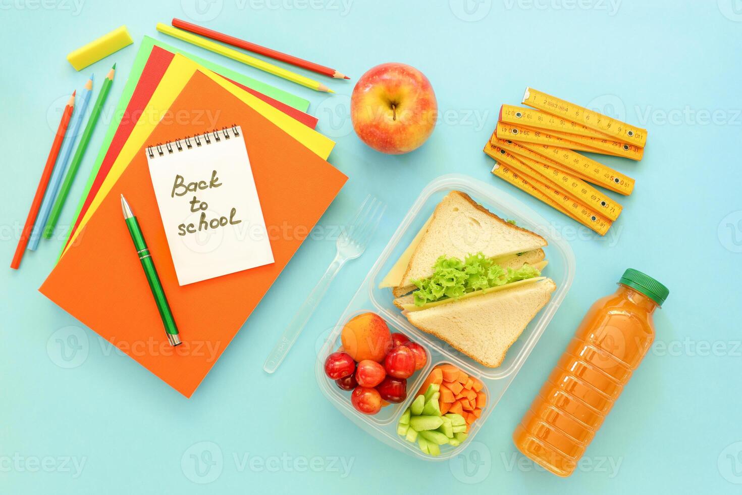 Lunchbox features sandwiches, fresh vegetables, fruits, along with stationery items like notebooks, pencils, and rulers on bright blue surface, preparing for school day. photo
