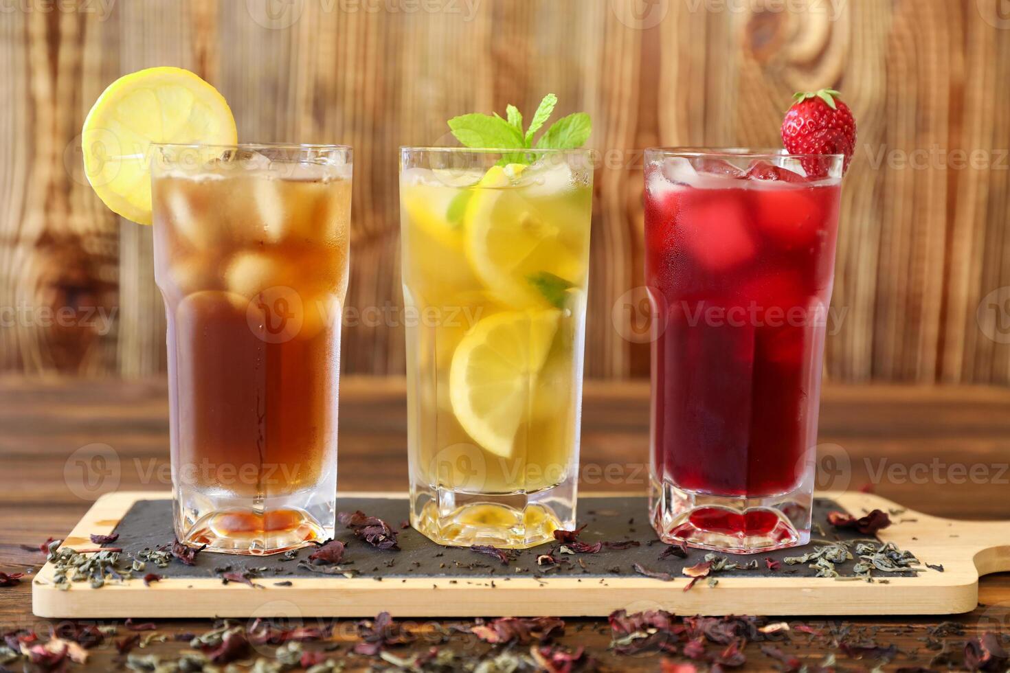Three tall glasses filled with refreshing drinks sit on wooden table. Iced tea features lemon slice, lemon drink contains fresh mint, and fruit punch showcases strawberries. photo