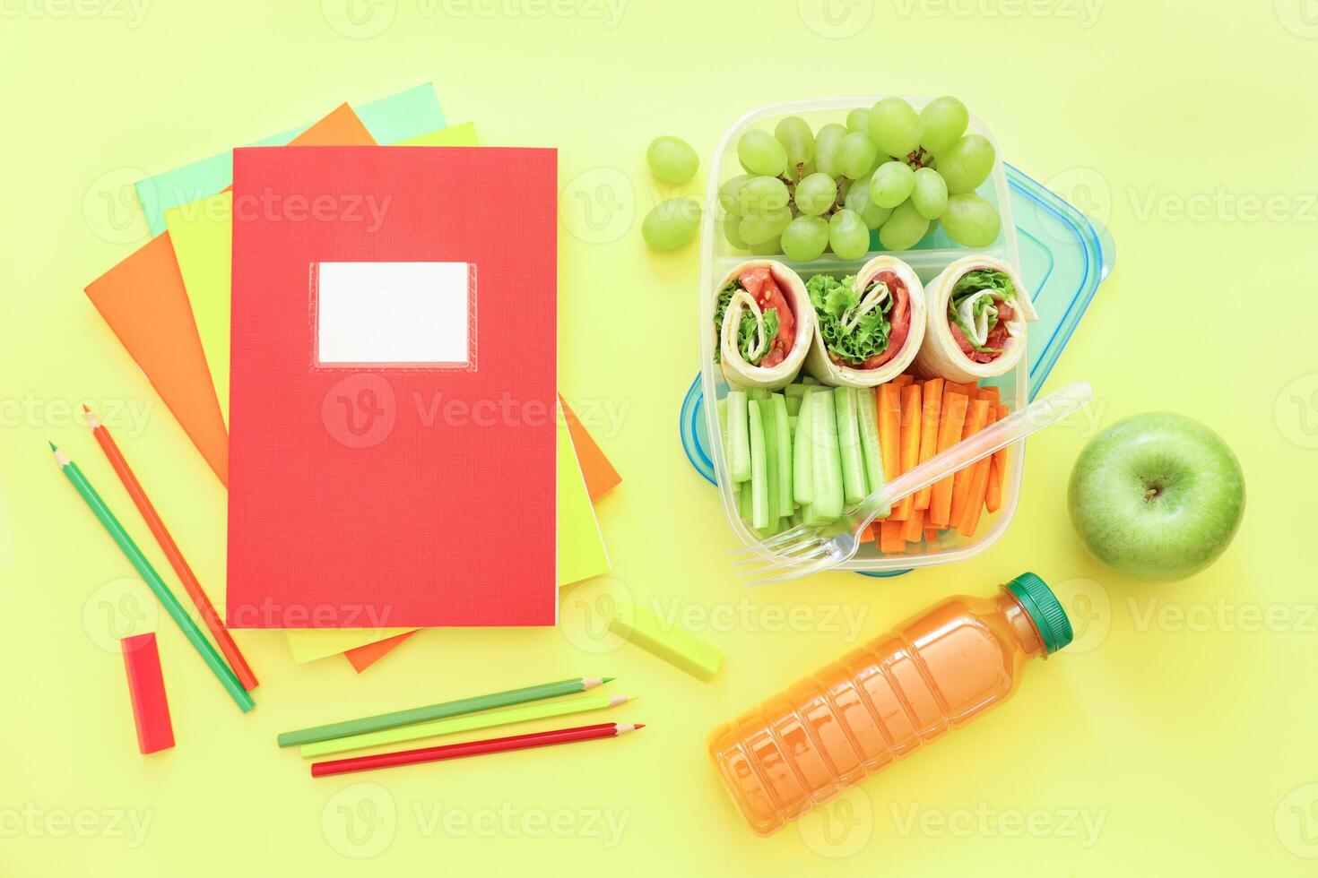 School supplies and lunch box with tasty rolls, cucumbers, carrots, grapes, apple, bottle of juice on light yellow background, back to school concept. Horizontal. Top view. photo