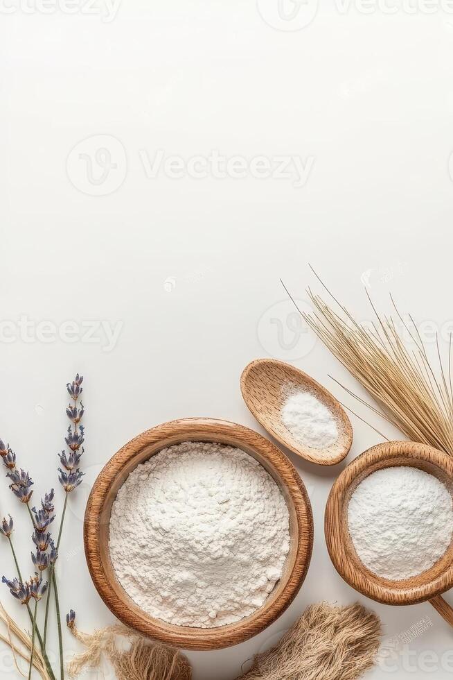 Wooden bowls contain white powder. Lavender flowers and dry wheat plants lie nearby on a plain surface with open space at top. photo