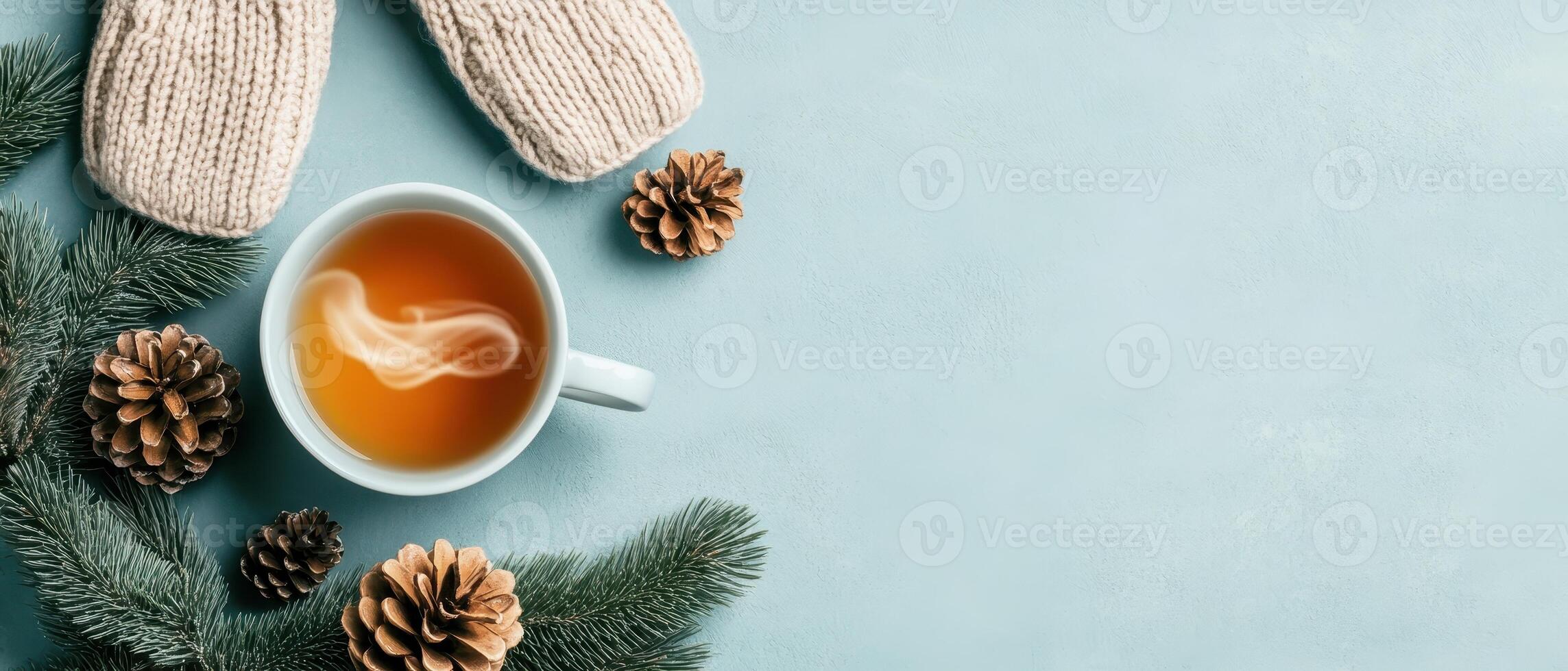 Warm drink steams in cup with mittens fir branches and pine cones on blue background. Winter cozy scene with copy space. photo