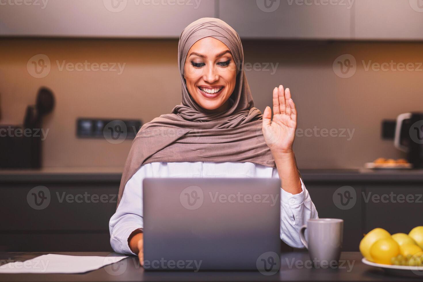 Muslim Lady At Laptop Making Call Waving Hello Sitting In Modern Kitchen At Home, Wearing Hijab. Arab Woman Using Computer Communicating Remotely. Female Dietitian Having Online Consultation. photo