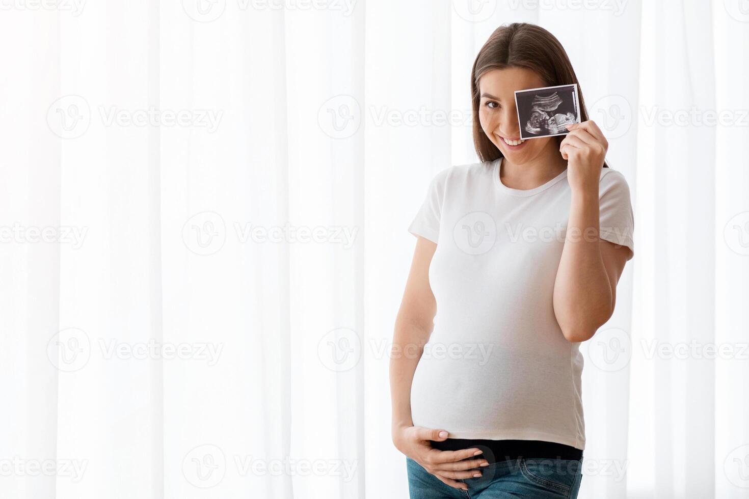 Happy Pregnant Woman With Ultrasound Scan Image Posing Near Window At Home, Cheerful Expecting Lady Embracing Her Belly And Smiling At Camera, Enjoying Healthy Pregnancy And Prenatal Time, Copy Space photo
