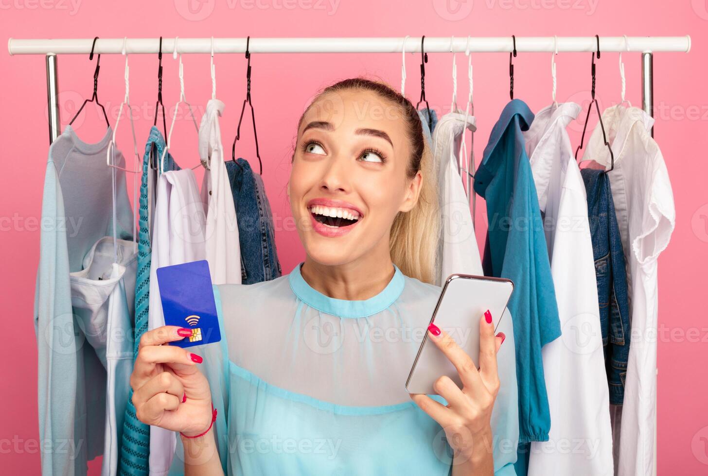 Retail And Purchase Concept. Excited stylish woman holding credit card and smartphone, looking up and thinking what to buy, ordering and buying clothes in online store via internet photo