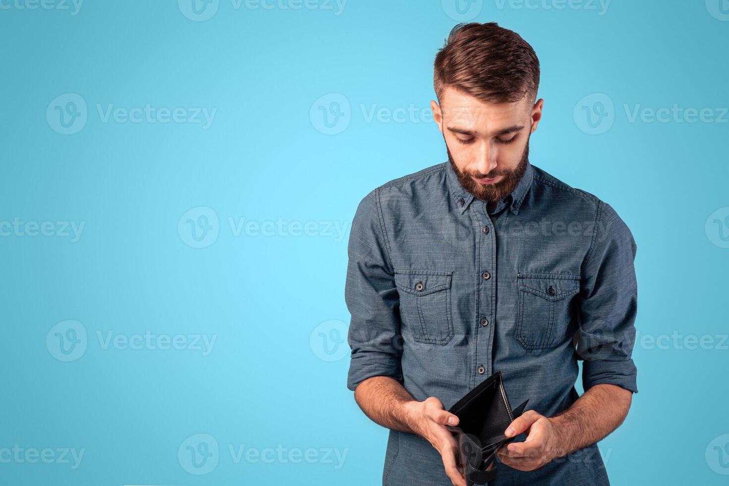 Poverty and unemployment concept. Desperate young man showing empty wallet with no money over blue studio background. Millennial guy losing his job or business during economic crisis photo
