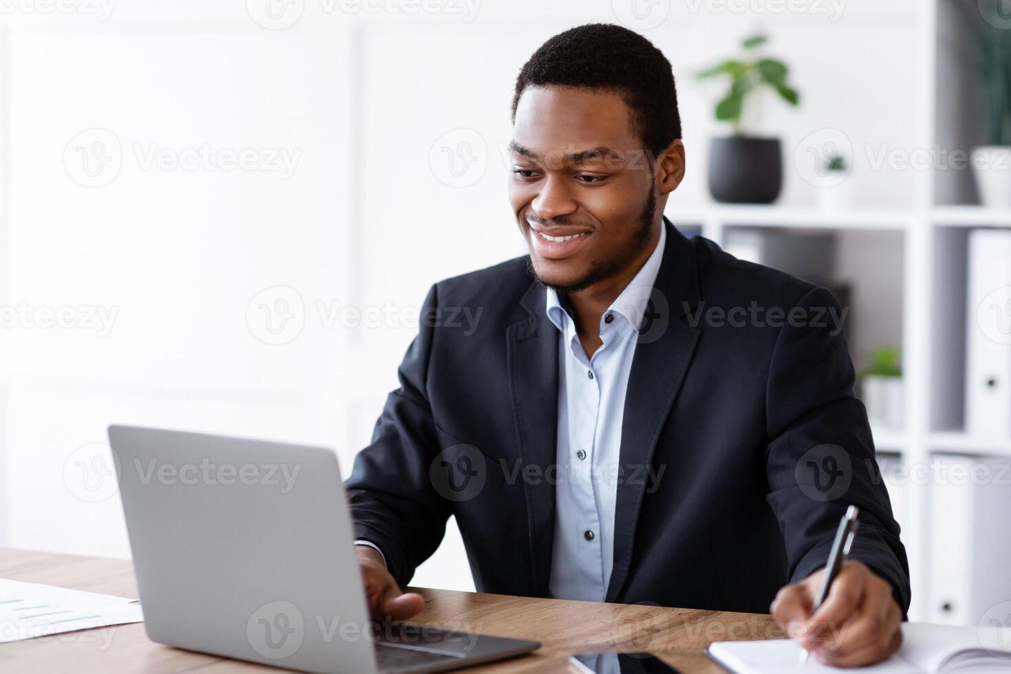 Cheerful young black businessman listening to webinar on laptop and taking notes, office interior, copy space. Smiling african american manager using laptop and writing something at notepad photo