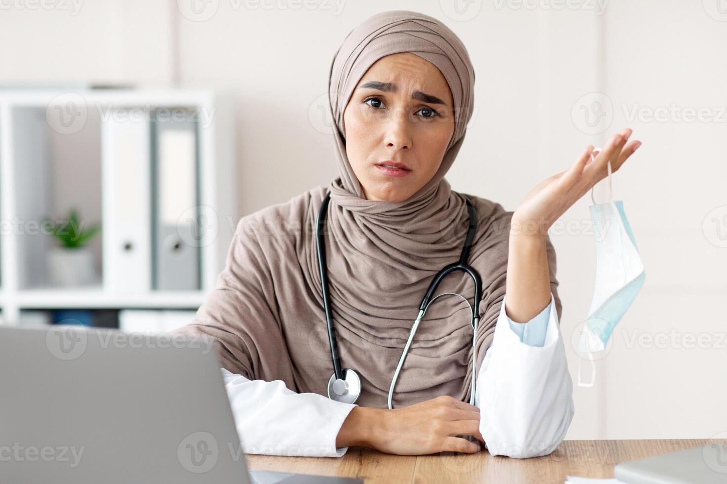 Annoyed muslim lady doctor removing protective face mask and complaining, feeling tired after difficult working day, sitting in front of laptop, copy space. Doctors and COVID-19 pandemic concept photo