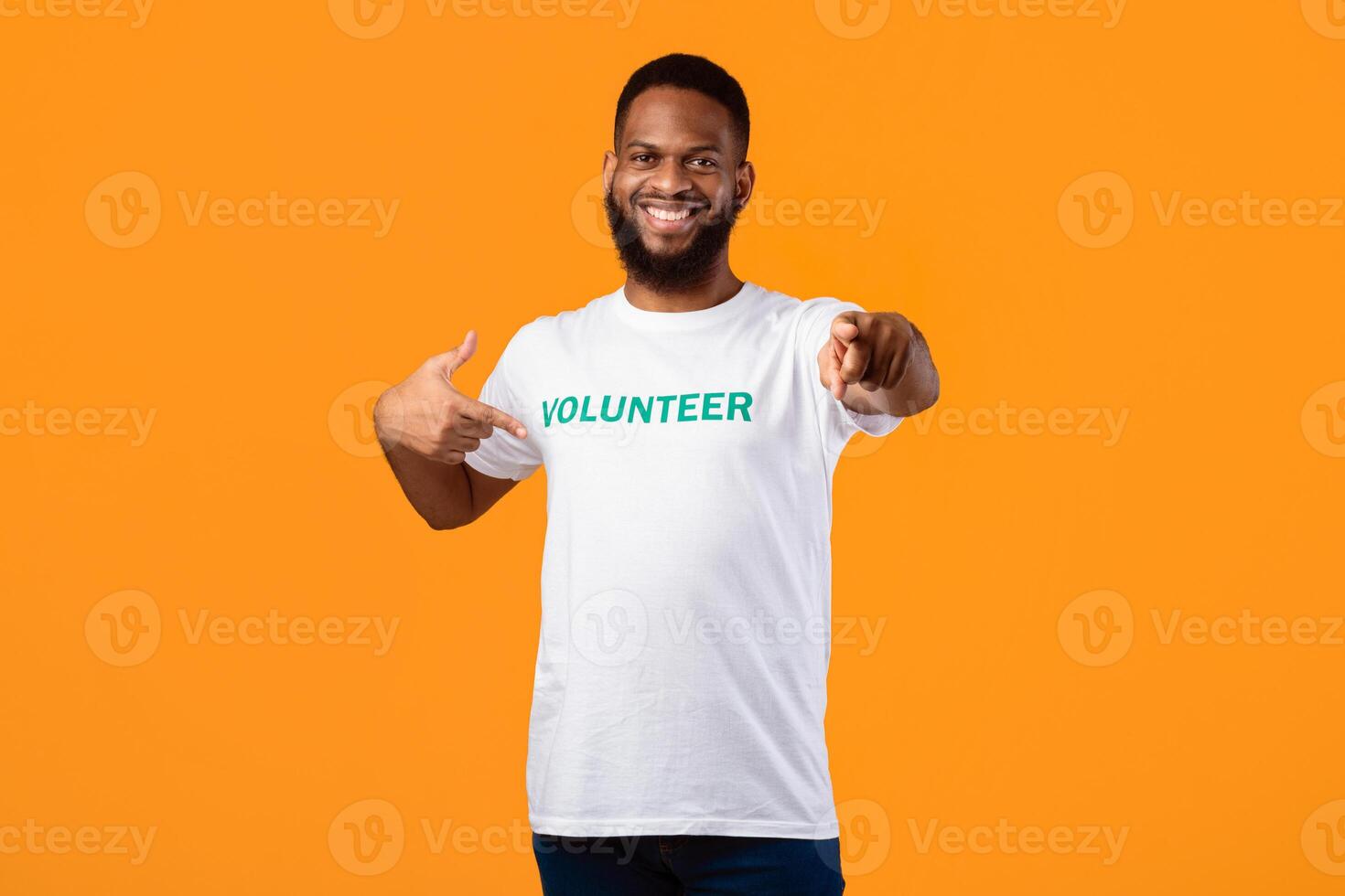 Happy black male volunteer activist pointing finger at his t-shirt and at camera posing on yellow studio background, inviting you to join volunteering career and work. You're next concept photo