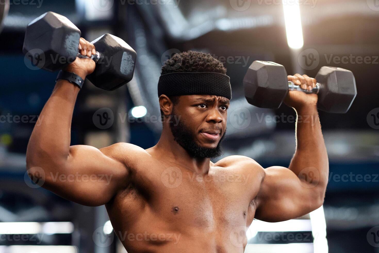 Confident african american shirtless man bodybuilder with muscular body exercising at gym, lifting up dumbbells, making work out for his biceps and back, looking at copy space photo