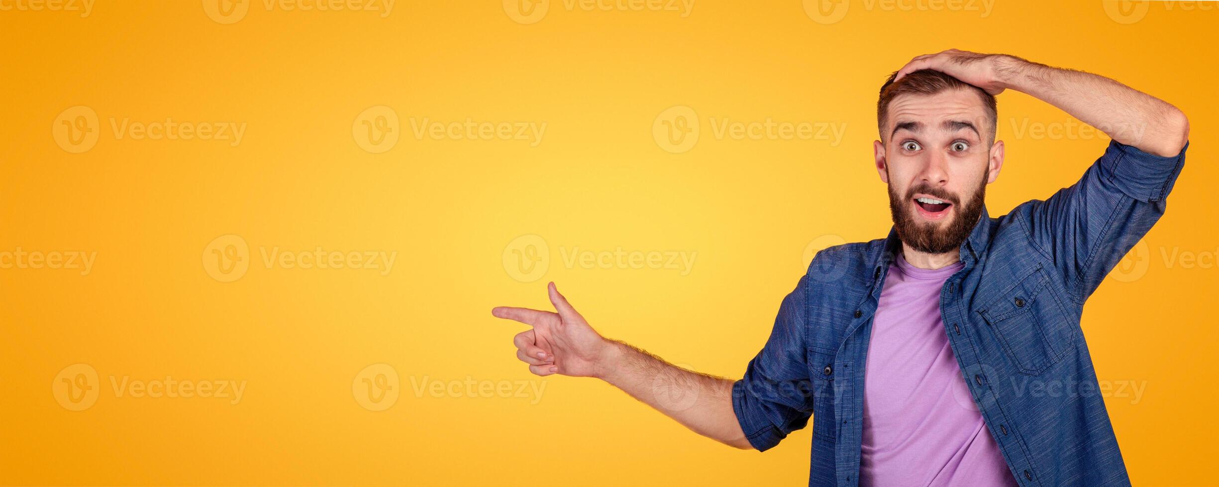 Shocked young man pointing aside at blank space on orange studio background, banner design. Millennial hipster guy holding his head in surprise, offering unbelievable sale or promo photo