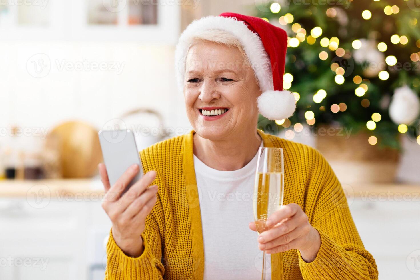 Positive granny in Santa hat having videocall on smartphone while sitting on festive kitchen, drinking champagne, copy space. Happy old lady celebrating New Year with her friends or family online photo