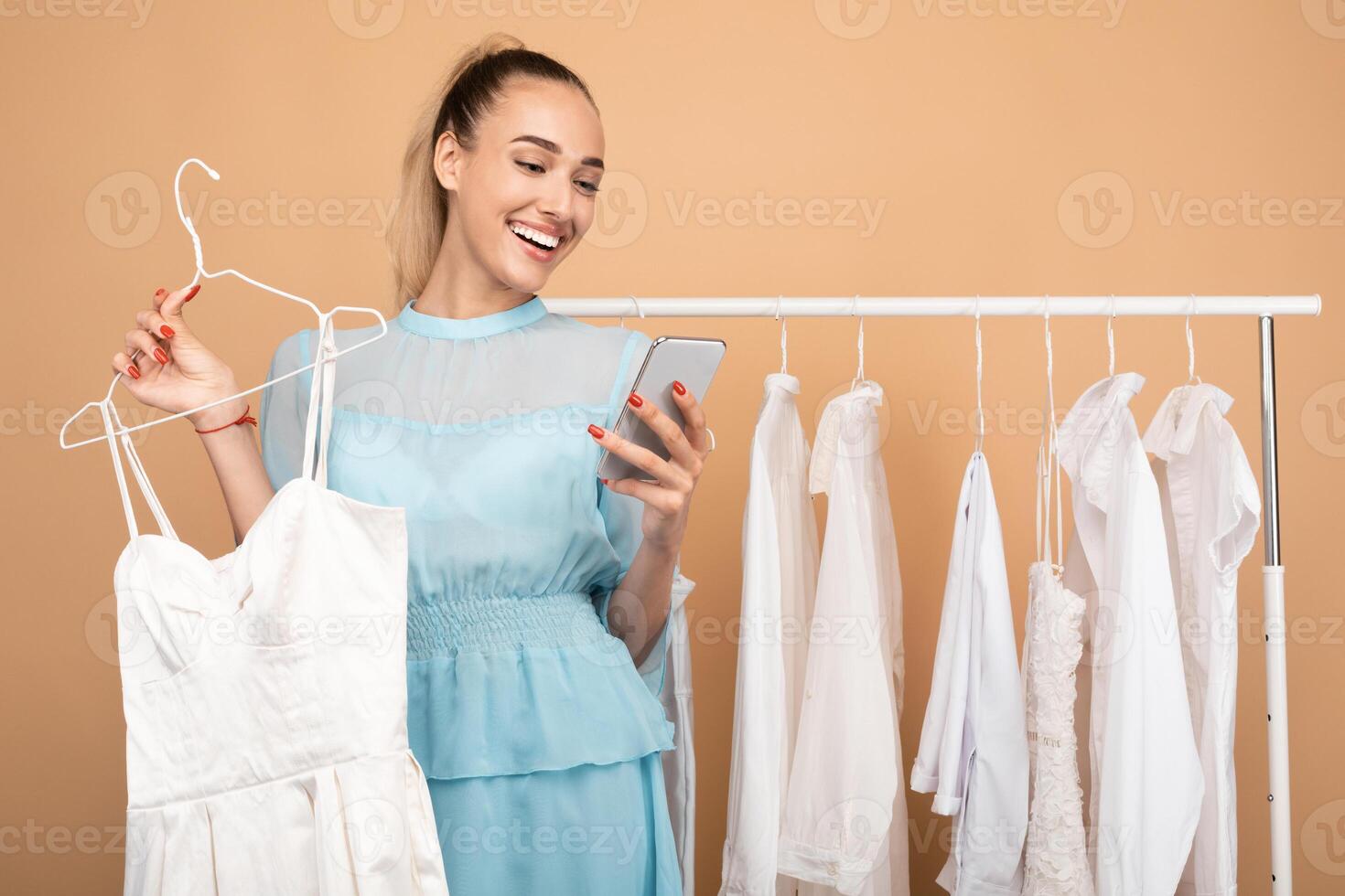Portrait of smiling young woman using her mobile phone, holding hanger with dress, chatting and sending photos to her friend, choosing outfit, standing near clothing rail at the studio