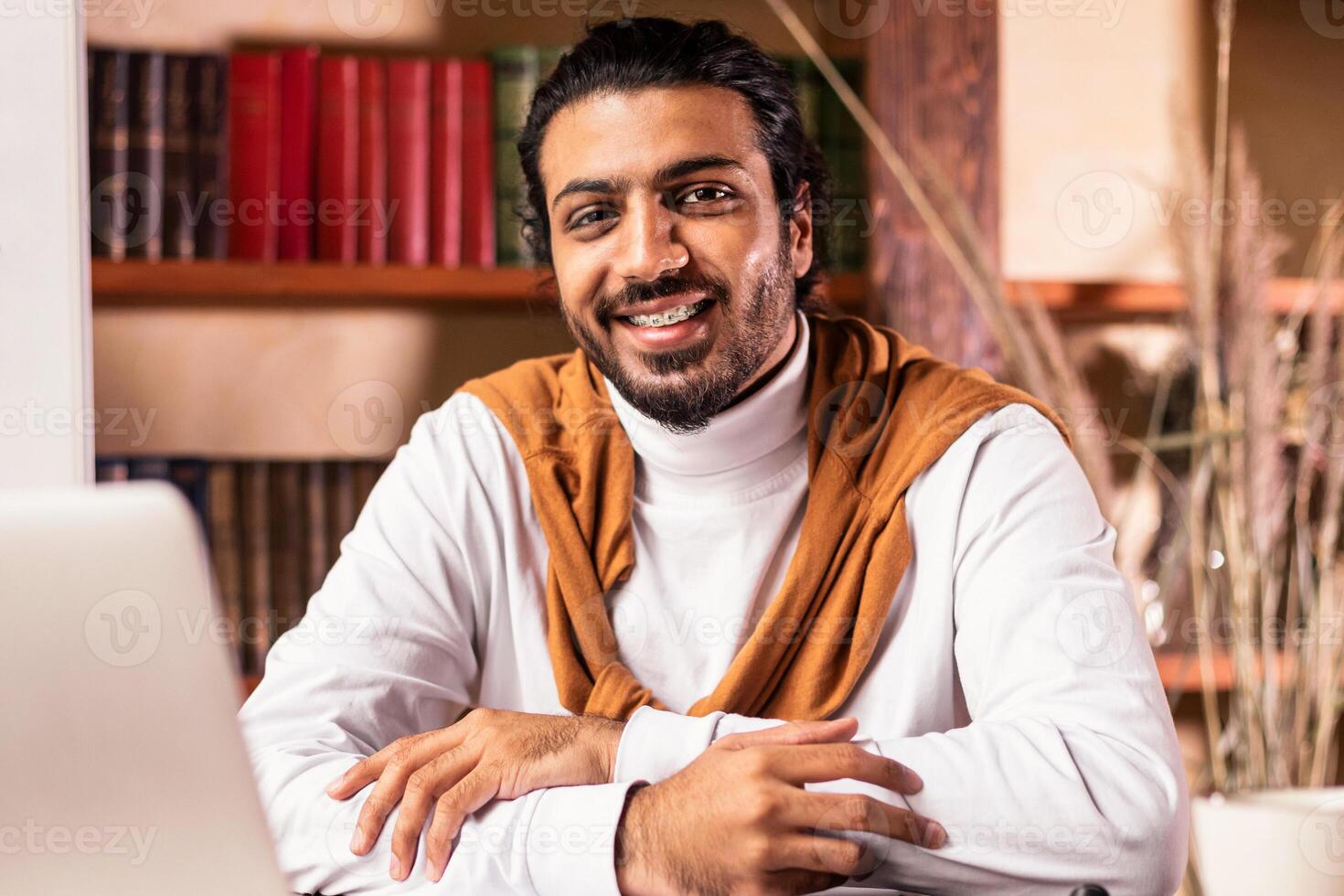 E-Teaching. Successful Indian Teacher Sitting At Laptop And Smiling To Camera Posing Teaching Online In University Library Indoors. Teacher's Portrait, Modern Education Concept photo