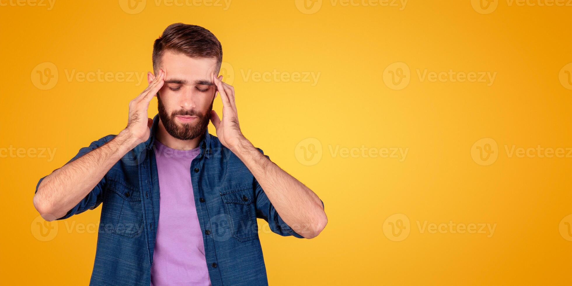 Young man suffering from headache, massaging his temples to ease pain on grey studio background. Millennial guy having high blood pressure, feeling stressed or tired, cannot solve problem photo