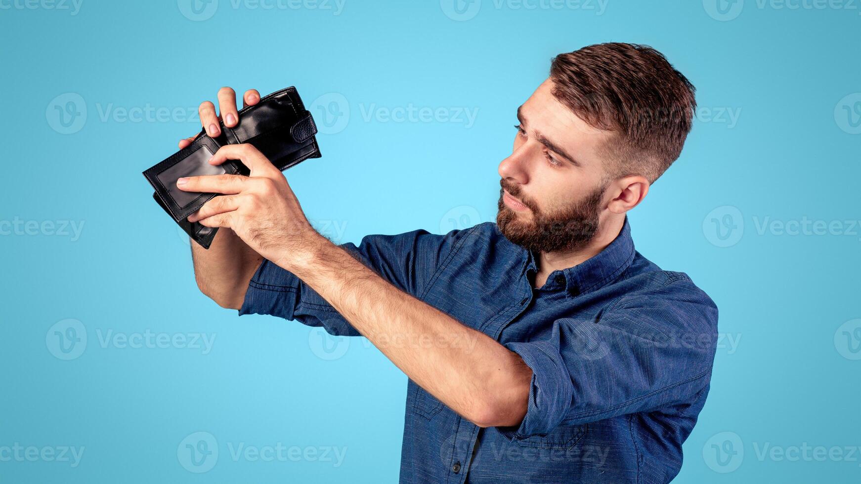 Bankruptcy, poverty and unemployment. Miserable young man looking into empty wallet without money over blue background. Moneyless millennial guy having financial problems during economic crisis photo