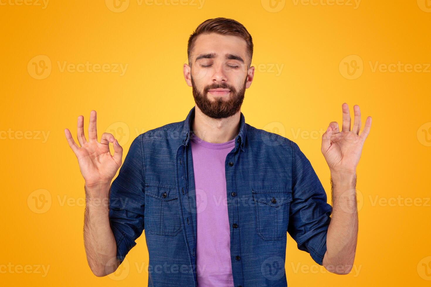 Stress relief concept. Handsome young man meditating with closed eyes, keeping calm on orange studio background. Millennial hipster guy practicing yoga, searching for inner balance photo