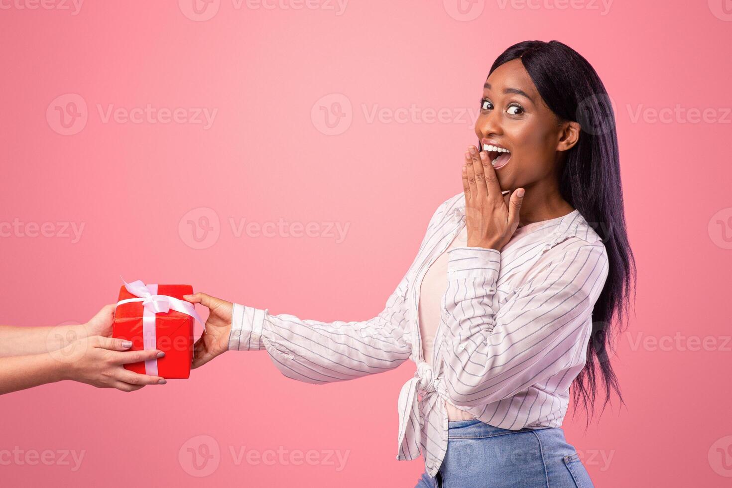 Surprised young lady receiving gift from female friend on pink studio background. Beautiful African American woman feeling excited to get wrapped present, celebrating holiday or special day photo