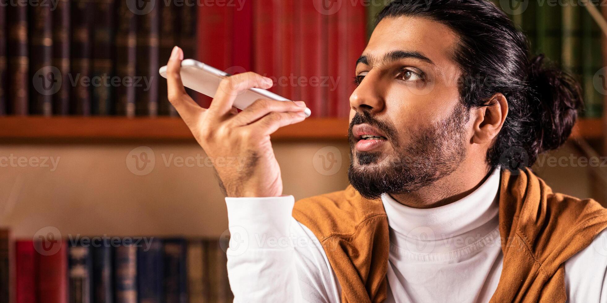 Indian Student Guy Using Voice Assistant On Smartphone Learning Sitting In University Library Indoors. Educational Application For Mobile Phone. Modern Education Concept. Selective Focus photo
