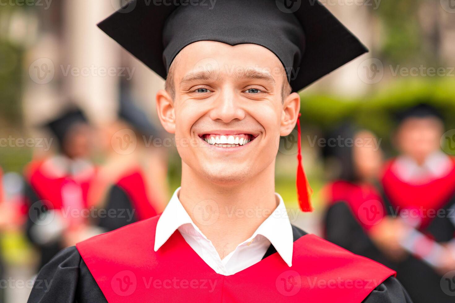 Handsome guy student posing in graduation costume, black and red robe and hat, cheerfully smiling at camera while having graduation ceremony at university campus, closeup portrait, copy space photo