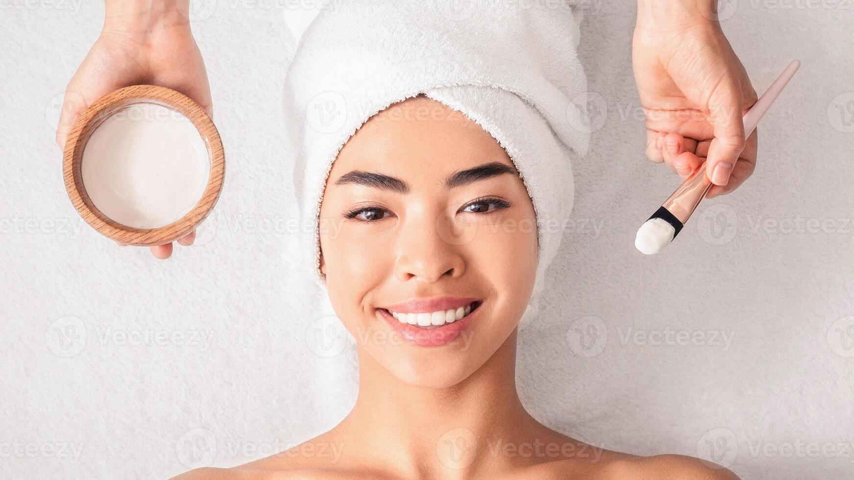 Facial Treatment. Top view of spa cosmetologist applying clay mask on asian woman's face, beautiful korean woman with towel on head lying on table enjoying skin care procedures in salon, panorama photo